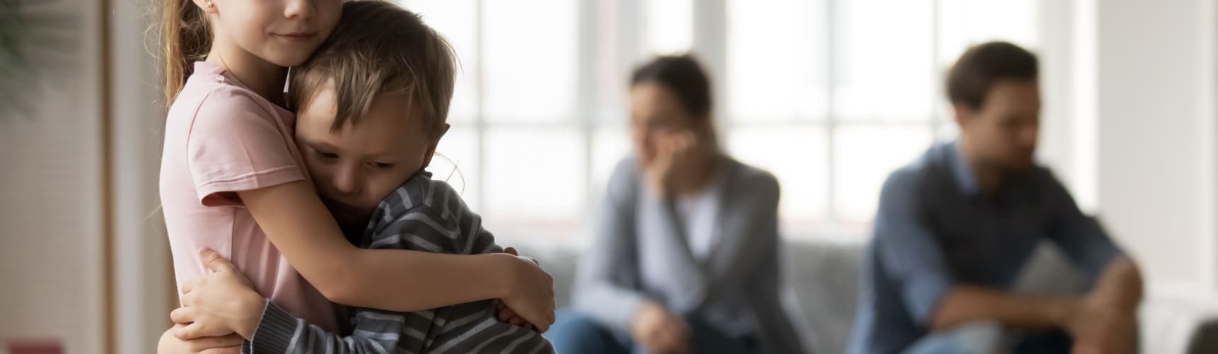 A pair of Children hugging, as their parents sit in the background